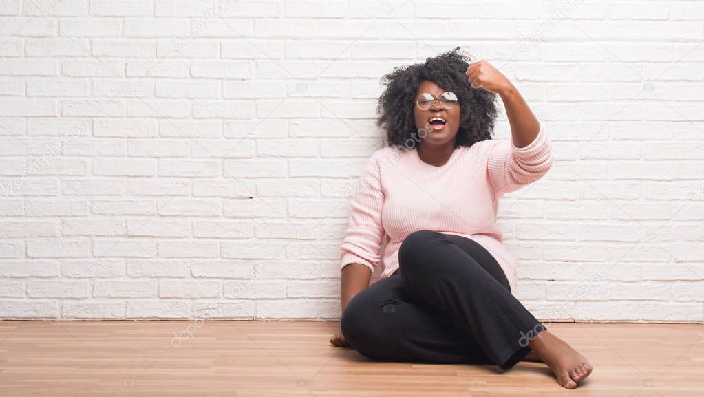 Young african american woman sitting on the floor at home angry and mad raising fist frustrated and furious while shouting with anger. Rage and aggressive concept.