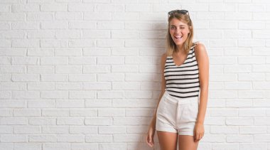 Genç güzel sarışın kadın seksi bir ifadeyle, neşeli ve mutlu yüzünü kameraya bakarak göz kırpıyor beyaz tuğla duvarın üzerinden.