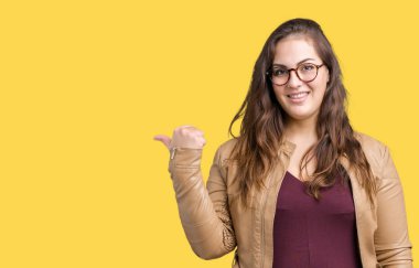 Seyir ve başparmak yanında yukarıyı mutlu yüz ile gülümseyen izole arka plan üzerinde elbise ve deri ceket giyen güzel artı boyutu genç kadın.