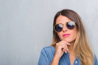 Grunge Genç Yetişkin kadın gri duvar çene düşünme hakkında soru, dalgın ifade üzerine el ile retro güneş gözlüğü takıyor. Düşünceli yüzüyle gülümsüyor. Şüphe kavramı.