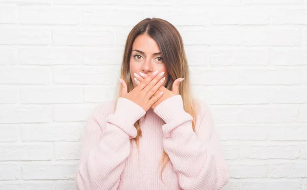 Young adult woman over white brick wall wearing winter outfit at home shocked covering mouth with hands for mistake. Secret concept.