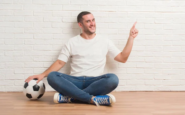非常に幸せな側の手と指で指すサッカー サッカー ボールを保持している白いレンガの壁に座っている若い白人男 — ストック写真