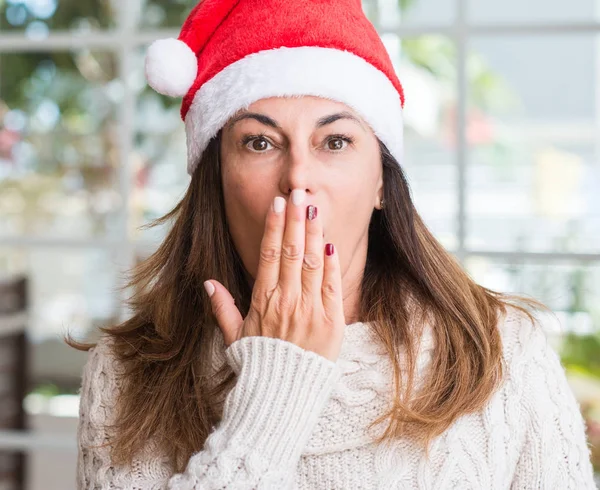 Middle aged woman wearing santa claus hat at home cover mouth with hand shocked with shame for mistake, expression of fear, scared in silence, secret concept