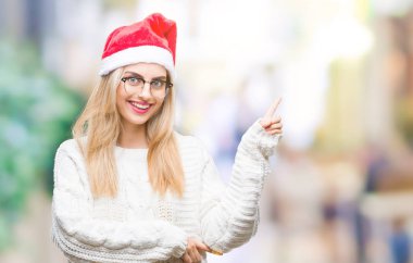 Kameraya bakarak yan el ve parmak ile işaret yüzünde kocaman bir gülümseme ile izole arka plan üzerinde Noel şapka giyen genç güzel sarışın kadın.