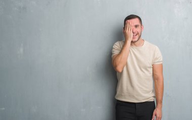 El yüz ve sürpriz duygu kendinden emin gülümseme ile tek gözü kaplayan gri grunge duvarın üzerinden beyaz delikanlı.