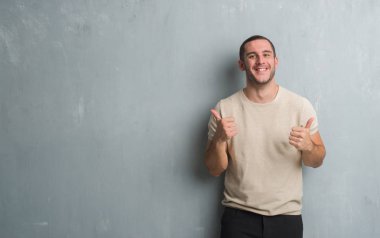 Beyaz delikanlı üzerinde gri grunge duvar başarı işareti başparmak gülümseyen ve mutlu el ile olumlu hareketi yapıyor. Neşeli bir ifadeyle, kazanan jest kameraya bakıyor.