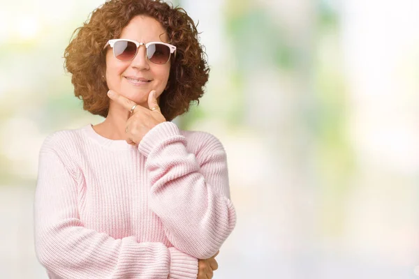Beautiful middle ager senior woman wearing pink sweater and sunglasses over isolated background looking confident at the camera with smile with crossed arms and hand raised on chin. Thinking positive.