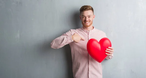 Young Redhead Man Grey Grunge Wall Holding Red Heart Surprise — Stock Photo, Image