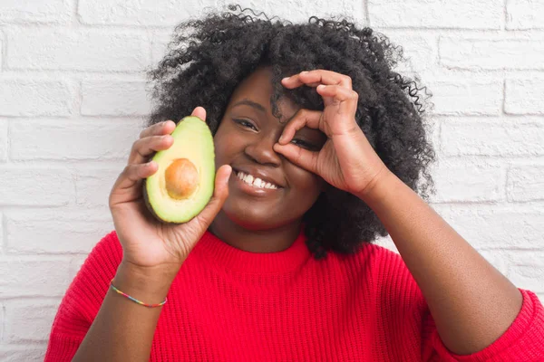 指の間から見て目に手で サインをして笑っている幸せそうな顔でアボカドを白いレンガ壁を食べる以上の若いアフリカ系アメリカ人女性 — ストック写真