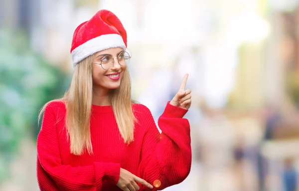 年轻美丽的金发女子戴着圣诞节帽子在孤立的背景与一个大微笑在脸上 指着手和手指在一边看着相机 — 图库照片