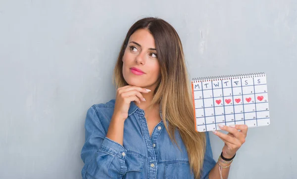Young adult woman over grey grunge wall showing period calendar serious face thinking about question, very confused idea