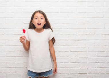 Kırmızı kalp lolipop şeker şok sürpriz yüzü, korkuyor ve korku ifadesi ile heyecanlı olan korkmuş beyaz tuğla duvarın üzerinden genç İspanyol çocuk