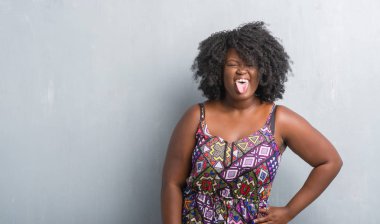 Genç Afro-Amerikan kadın dilini renkli elbise gri grunge duvarın üzerinden out komik ifade ile mutlu. Duygu kavramı.