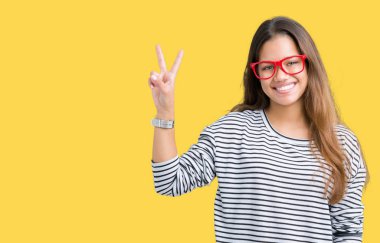 Giyen genç güzel esmer kadın kazak ve kırmızı gözlük mutlu yüz zafer işareti yaparken kameraya göz kırpıyor ile gülümseyen izole arka plan üzerinde çizgili. İki numaralı.