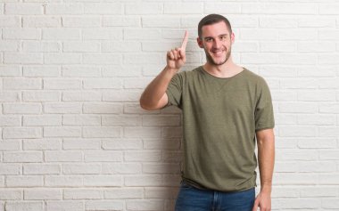 Genç beyaz adam ve parmak gülümseyen süre bir numara ile kendinden emin ve mutlu yukarıyı gösteren beyaz tuğla duvarın üzerinden ayakta.