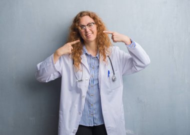 Genç redhead doktor kadın kendinden emin gösterilen ve işaret parmakları diş ve ağız ile gülümseyen bir palto giyiyor gri grunge duvarın üzerinden. Sağlık kavramı.