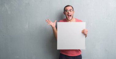 Boş bir holding gri grunge duvarın üzerinden beyaz delikanlı çok mutlu ve heyecanlı, kazanan ifade büyük gülümseme ile çığlık zaferini kutluyor afiş ve ellerini kaldırdı