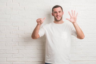 Beyaz delikanlı gösterilen ve parmakları ile işaret beyaz tuğla duvarın üzerinden ayakta 6 kendinden emin ve mutlu gülümseyerek iken numara.
