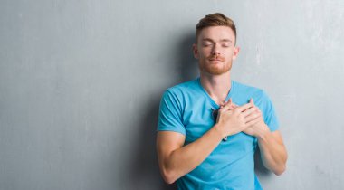 Genç Kızıl saçlı adam gri grunge duvar kapalı gözler ile göğüs ellerinde ve yüzünde minnettar jest ile gülümseyen giyiyor rahat kıyafet. Sağlık kavramı.