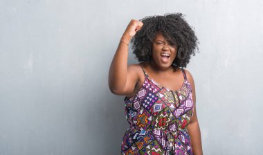 Gri grunge duvar öfkeli ve sinirli sinirli ve öfke ile bağırarak süre öfkeli yumruk yükselterek renkli elbise giyen genç Afro-Amerikan kadın. Öfke ve agresif kavramı.
