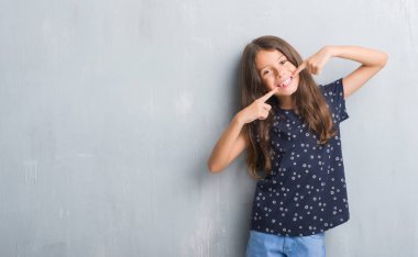 Grunge gri duvar emin gösterilen ve işaret parmakları diş ve ağız ile gülümseyen genç İspanyol çocuk. Sağlık kavramı.