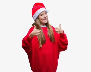 El ile pozitif jest yapıyor izole arka plan başarı işareti üzerinde Noel şapka giyen genç güzel kız gülümseyerek ve mutlu yaşasın. Neşeli bir ifadeyle, kazanan jest kameraya bakıyor.