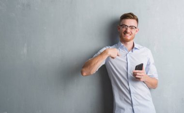 Genç Kızıl saçlı adam kendisi için sürpriz yüz işaret parmağı ile smartphone kullanarak ileti gönderme gri grunge duvarın üzerinden