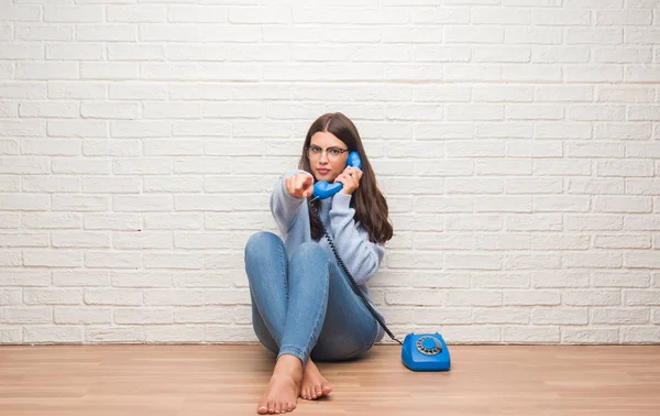Young brunette woman sitting on the floor calling on vintage telephone pointing with finger to the camera and to you, hand sign, positive and confident gesture from the front