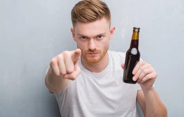 年轻的红发男子在灰色的格格墙上喝啤酒瓶用手指指向相机和你 手的标志 积极和自信的手势从前面 — 图库照片