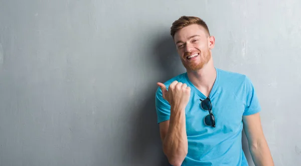 Young redhead man over grey grunge wall wearing casual outfit smiling with happy face looking and pointing to the side with thumb up.