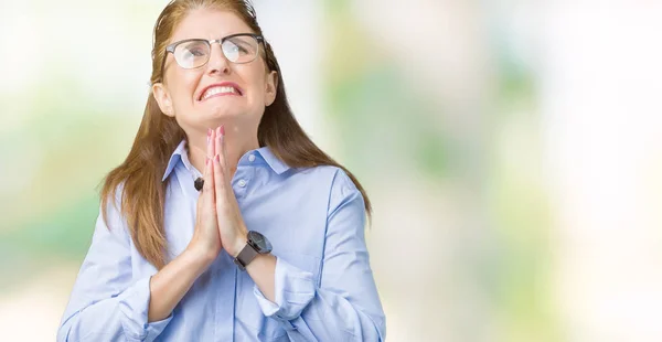 Beautiful middle age mature business woman wearing glasses over isolated background begging and praying with hands together with hope expression on face very emotional and worried. Asking for forgiveness. Religion concept.