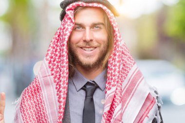 Genç yakışıklı Arap adam uzun saçlı keffiyeh gösterilen ve parmakları ile işaret izole arka plan üzerinde giyen ikincisi ise kendinden emin ve mutlu gülümseyerek.