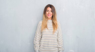 Grunge gri duvar yüzündeki mutlu ve güzel bir gülümseme ile kış kazak giyen genç yetişkin kadın. Şanslı kişi.