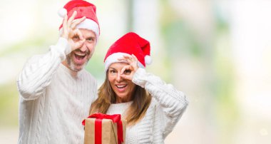 Noel şapka ve hediye ile mutlu yüz el parmaklarının arasından seyir göz üzerinde ok işaretiyle yapıyor gülümseyen izole arka plan üzerinde tutan orta yaş İspanyol çift