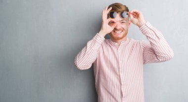 Genç Kızıl saçlı adam mutlu yüz el parmaklarının arasından seyir göz üzerinde ok işaretiyle yapıyor gülümseyen retro güneş gözlüğü takıyor gri grunge duvarın üzerinden