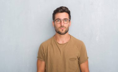 Yakışıklı genç adam gözlüklü ciddi ifade ile yüzünde gri grunge duvarın üzerinden. Basit ve doğal kameraya bakıyor.
