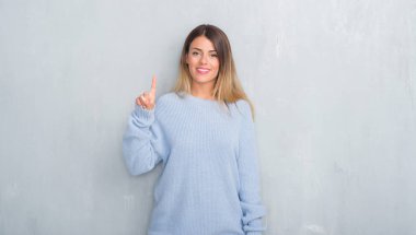 Gri grunge duvar kış kıyafeti gösterme ve kendinden emin ve mutlu gülümseyen bir parmak ile işaret eden oktan yukarı giyen genç yetişkin kadın.