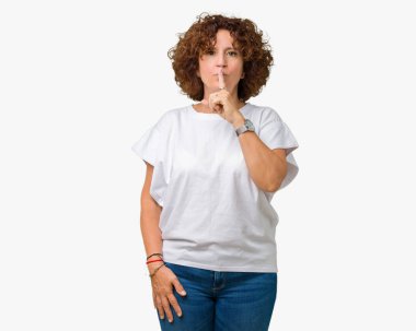 Dudaklarında parmak ile sessiz olmanı izole arka plan üzerinde beyaz t-shirt giyen güzel orta ager kadın kıdemli. Sessizlik ve gizli kavramı.