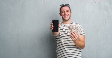 Beyaz delikanlı dolar ve smarphone mutlu ve güzel bir gülümseme ile yüz tutan gri grunge duvarın üzerinden. Şanslı kişi.