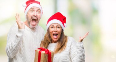 Orta yaş İspanyol çift giyen Noel şapka ve holding hediye üzerinde kocaman bir gülümseme ile çığlık zaferini kutluyor ve ellerini kaldırdı arka plan çok mutlu ve heyecanlı, kazanan ifade izole