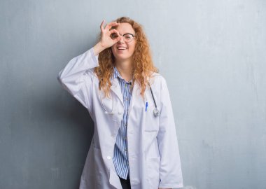 Genç redhead doktor kadın gülümsüyor, parmakları ile mutlu yüz üzerinden seyir göz el ile ok jest yapıyor bir palto giyiyor gri grunge duvarın üzerinden.