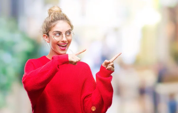Young beautiful blonde woman wearing red sweater and glasses over isolated background smiling and looking at the camera pointing with two hands and fingers to the side.