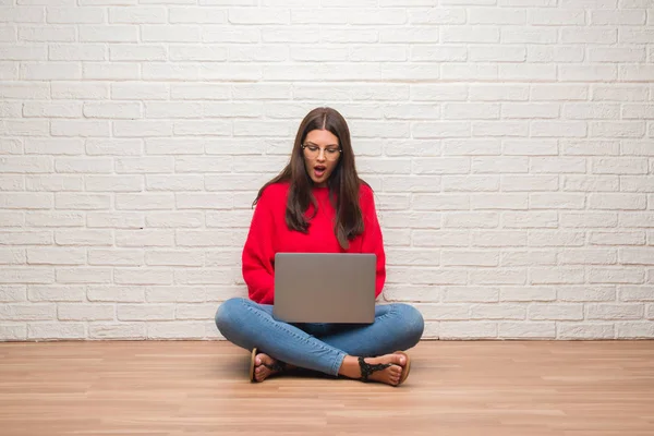年轻的黑发女子坐在地板上 坐在白色砖墙上 用电脑电脑吓得目瞪口呆 害怕和兴奋的恐惧表情 — 图库照片