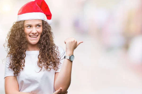 年轻的黑发女孩在孤立的背景上戴着圣诞帽微笑着 愉快的脸看着 用拇指指着一边 — 图库照片
