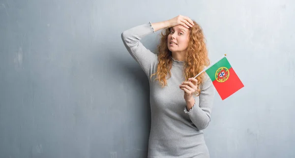 年轻的红发女子在灰色的沙哑墙上拿着葡萄牙的国旗 用手在头上强调 震惊与耻辱和惊喜的脸 愤怒和沮丧 因错误而恐惧和不安 — 图库照片