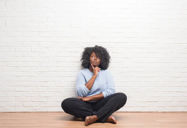 若いアフリカ系アメリカ人の女性はあごを考えて質問 物思いにふける式の手で家の床の上に座って 思いやりのある顔をして笑っています 疑いのコンセプト — ストック写真