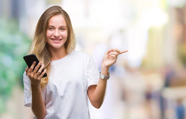 年轻美丽的金发女子使用智能手机在孤立的背景非常愉快的指向与手和手指的一侧 — 图库照片