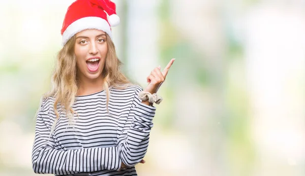 美しい若いブロンドの女性の顔に大きな笑みを浮かべて隔離された背景にクリスマスの帽子を着てカメラを見て側の手と指で指す — ストック写真