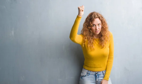 Young redhead woman over grey grunge wall angry and mad raising fist frustrated and furious while shouting with anger. Rage and aggressive concept.