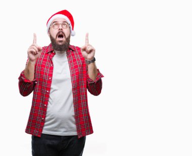 Noel şapka izole arka plan üzerinde beyaz delikanlı hayrete ve yukarıya bakıyor ve parmakları ile işaret ve silah kaldırdı şaşırttı.